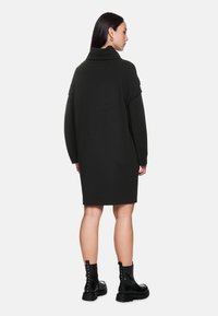 Schwarzes, oversized Strickkleid mit hohem Kragen, gerippten Details an den Ärmeln und geradem Schnitt, kombiniert mit schwarzen Stiefeletten.
