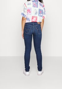 Jeans skinny blu scuro con tasche posteriori e cuciture caratteristiche, abbinati a una camicia a maniche corte con motivo in colori chiari.