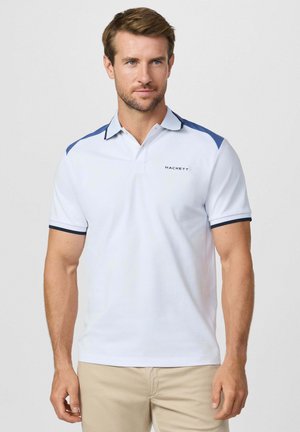 Man met een wit poloshirt met blauwe schouderpanelen en beige broek, staand tegen een effen lichte achtergrond.