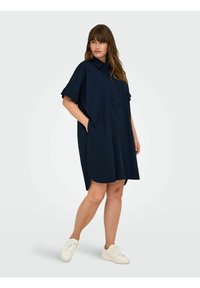 Rochie cămașă bleumarin, cu mâneci scurte, deschisă la nasturi și buzunare laterale. Prezintă o croială lejeră și un tiv rotunjit. Poartă adidași albi.