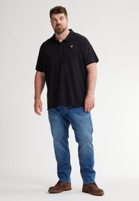 Musta polo-paita, jossa on brodeerattu logo, yhdistettynä sinisiin farkkuihin ja ruskeisiin kenkiin. Lyhyet hihat, rento malli ja sileä pinta.