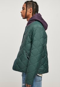 Chaqueta acolchada de color verde oscuro con textura esponjosa, que cuenta con cierre a presión, bolsillos laterales y una sudadera con capucha morada debajo.