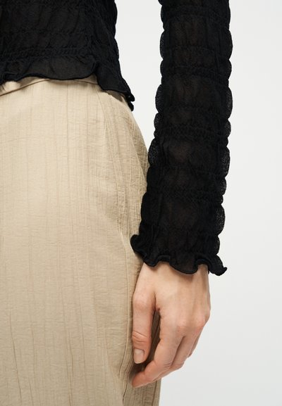 Χέρι με ανοιχτό χρώμα δέρματος, ακουμπισμένο δίπλα σε μπεζ υφασμάτινη φούστα με υφή και μαύρο μακρυμάνικο τοπ με δαντελωτό μοτίβο.