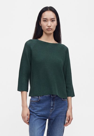 Femme aux longs cheveux foncés portant un haut en tricot texturé vert foncé à manches trois-quarts et un jean bleu, debout devant un fond blanc uni.