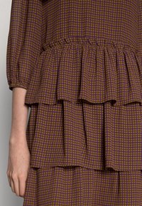 Braun kariertes Kleid mit gestuften Rüschen, aus einem strukturierten Stoff, elastischem Bund und lila Akzenten im Muster.