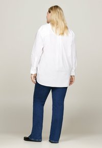 Žena so stredne dlhými blond vlasmi, oblečená v bielej košeli s dlhým rukávom, tmavomodrých džínsoch a čiernych topánkach, stojaca s chrbtom otočeným dopredu.