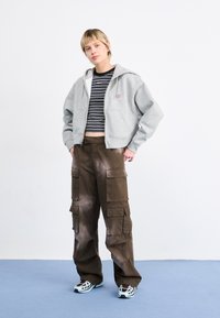 Felpa grigia corta con zip frontale e tasche, abbinata a pantaloni cargo marroni con multiple tasche e una vestibilità comoda.