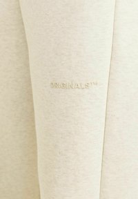 Lys beige sweatshirtstof med en glat tekstur, prydet med det præget "ORIGINALS™" logo på ærmet. Enkel, ren design.