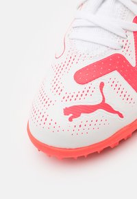 Scarpa sportiva bianca con accenti rosa. Presenta una superficie testurizzata, motivi a pois e un logo Puma ben visibile. Suola in gomma con tacchetti.