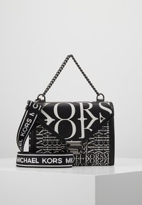Borsa Michael Kors in bianco e nero con grande stampa del logo, manico a catena e tracolla rimovibile con nome del marchio su superficie bianca.