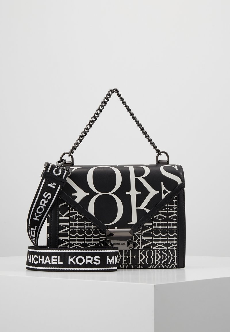 Borsa Michael Kors in bianco e nero con grande stampa del logo, manico a catena e tracolla rimovibile con nome del marchio su superficie bianca.