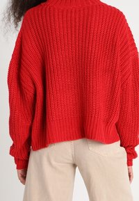Maglione rosso lavorato a maglia con un collo alto e una vestibilità over size, caratterizzato da un motivo testurizzato e polsini a costine, abbinato a pantaloni beige.