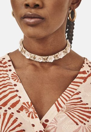 Collar choker floral blanco con acentos de cuentas, que presenta una superficie lisa y un diseño estructurado. Se lleva con una prenda de escote en V con patrón.