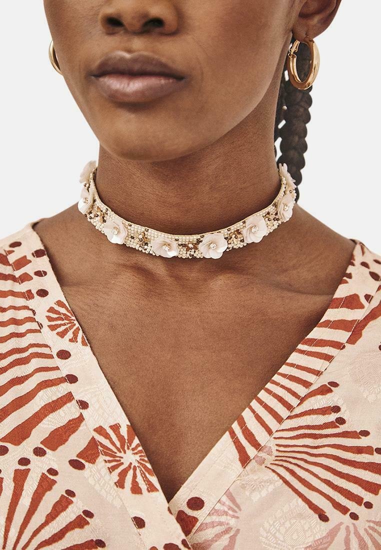 Collar choker floral blanco con acentos de cuentas, que presenta una superficie lisa y un diseño estructurado. Se lleva con una prenda de escote en V con patrón.