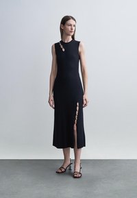 Moteris dėvi juodą midi suknelę be rankovių, su metaliniais žiedais ant peties ir šlaunies skeltuku, derinančią su juodais basutėmis su dirželiais.