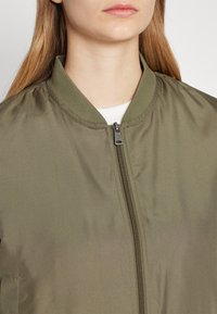 Olivově zelená bomber bunda s hladkým povrchem, žebrovaným límcem a centrálním zipem. Vyznačuje se uzavřeným designem manžet a minimálním zdobením.