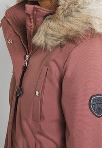 Chaqueta parka aislante rosa, con capucha de piel sintética, cierres de cremallera y botones, con bolsillos frontales y un parche con el logo en la manga.