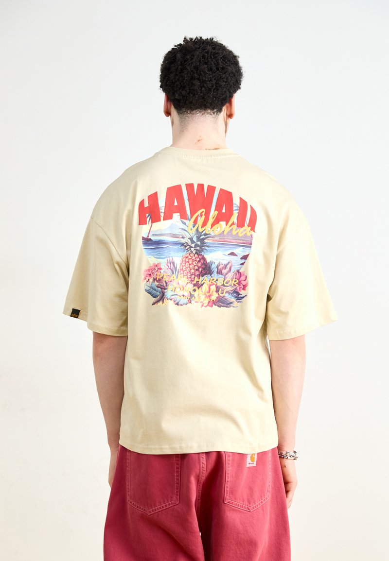 Beige kortermet t-skjorte med et fargerikt Hawaii-motiv på baksiden, som har ananas, havscene og tropiske elementer.