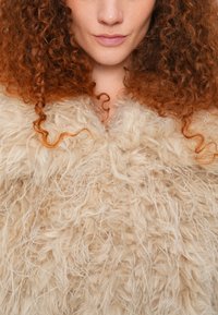 Μπεζ εξωτερικό ένδυμα από συνθετική γούνα με χνουδρή υφή και χαρακτηριστική όψη. Μαλακές, μακριές ίνες δημιουργούν ογκώδη εμφάνιση. Χωρίς ορατά κουμπώματα.