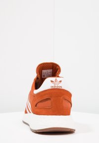 Orange mockasinsneakers med vita textilaccents och tre-stripesdesign. Har vadderad krage och en strukturerad vit sula.