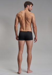 Boxer shorts noirs avec une texture lisse et une ceinture logo orange. Le modèle est montré de dos, debout pieds nus.
