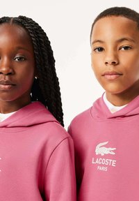 Due bambini indossano felpe coordinati di colore rosa con il logo bianco che recita "LACOSTE PARIS". Il tessuto sembra morbido e presenta una texture liscia.