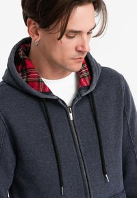 Felpa zip con cappuccio di colore blu navy realizzata in tessuto morbido, caratterizzata da una fodera in flanella a quadri rossi e neri nel cappuccio e da lacci neri.