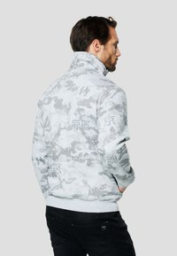 Felpa grigia mimetica con zip e collo alto, caratterizzata da testo e motivi neri a contrasto. Tessuto morbido e orlo a coste.