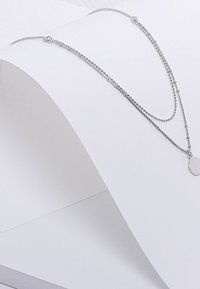 Zilveren gelaagde ketting met twee kettingen van verschillende lengtes, waarvan er één kleine ronde kralen en een platte ronde hanger heeft. Glad van textuur.