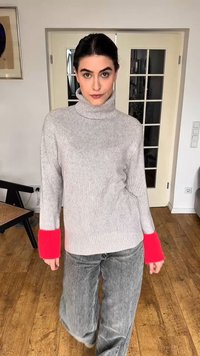 Femme aux cheveux foncés attachés, portant un pull gris à col roulé avec des poignets rose vif et un jean gris ample, debout à l'intérieur sur un sol en bois.