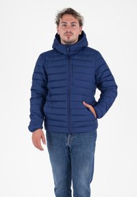 Jonge man in een marineblauwe gewatteerde jas met capuchon en een rits aan de voorkant, staande met één hand in zijn zak tegen een witte achtergrond.