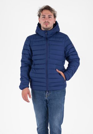 Jonge man in een marineblauwe gewatteerde jas met capuchon en een rits aan de voorkant, staande met één hand in zijn zak tegen een witte achtergrond.