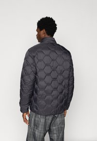 Man wearing a black quilted jacket with hexagonal stitching, facing away, paired with loose gray plaid trousers.