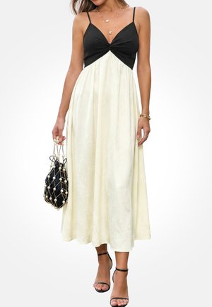 Vrouw draagt een zwart-witte mouwloze midijlange jurk met dunne bandjes, houdt een zwart netzakje met parelversiering vast en draagt zwarte hakken met enkelbandje.