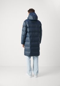Marineblauer Steppmantel mit Kapuze, mit gesteppten Bereichen, Logo-Patch am Ärmel, kombiniert mit hellblauen Jeans und weißen Schuhen.