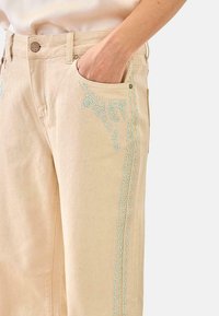 Primer plano de pantalones beige con bordado floral azul claro a lo largo de la costura lateral y el borde del bolsillo, mano descansando dentro del bolsillo delantero.