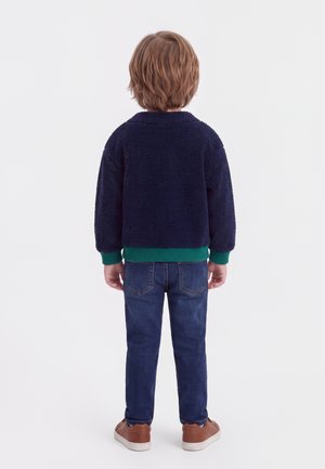 Enfant aux cheveux châtain clair portant une veste polaire bleu marine, un jean bleu et des chaussures marron, debout de dos sur fond blanc.