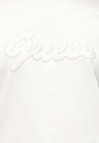 Λευκό βαμβακερό t-shirt με λαμπερό, υφασμάτινο λογότυπο "Guess" από μικρές χάντρες, τοποθετημένο κεντρικά στο μπροστινό μέρος.