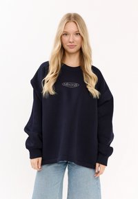 Granatowy, oversize'owy sweatshirt z prążkowanym dekoltem, opuszczonymi ramionami oraz subtelnie wyhaftowanym logiem "MIRONS". W połączeniu z jasnymi dżinsami.