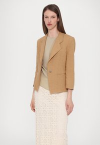 Lys brun lin blazer med en enkel knapp, spisse lapler og trekvart ermer, kombinert med en beige topp og et hvitt heklett skjørt.