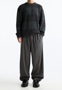 Personne portant un pull ample à rayures foncées, un pantalon gris large, des chaussures noires épaisses, debout devant un fond blanc.