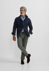 Chaqueta ligera navy con dos bolsillos en el pecho, combinada con una camisa azul clara, pantalones cargo oliva y zapatos de ante marrón.