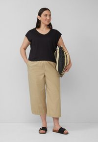 Zwarte v-hals top met korte mouwen, beige cropped broek en zwarte sandalen. Het model houdt een gestreepte geweven tas vast. Gladde texturen overal.
