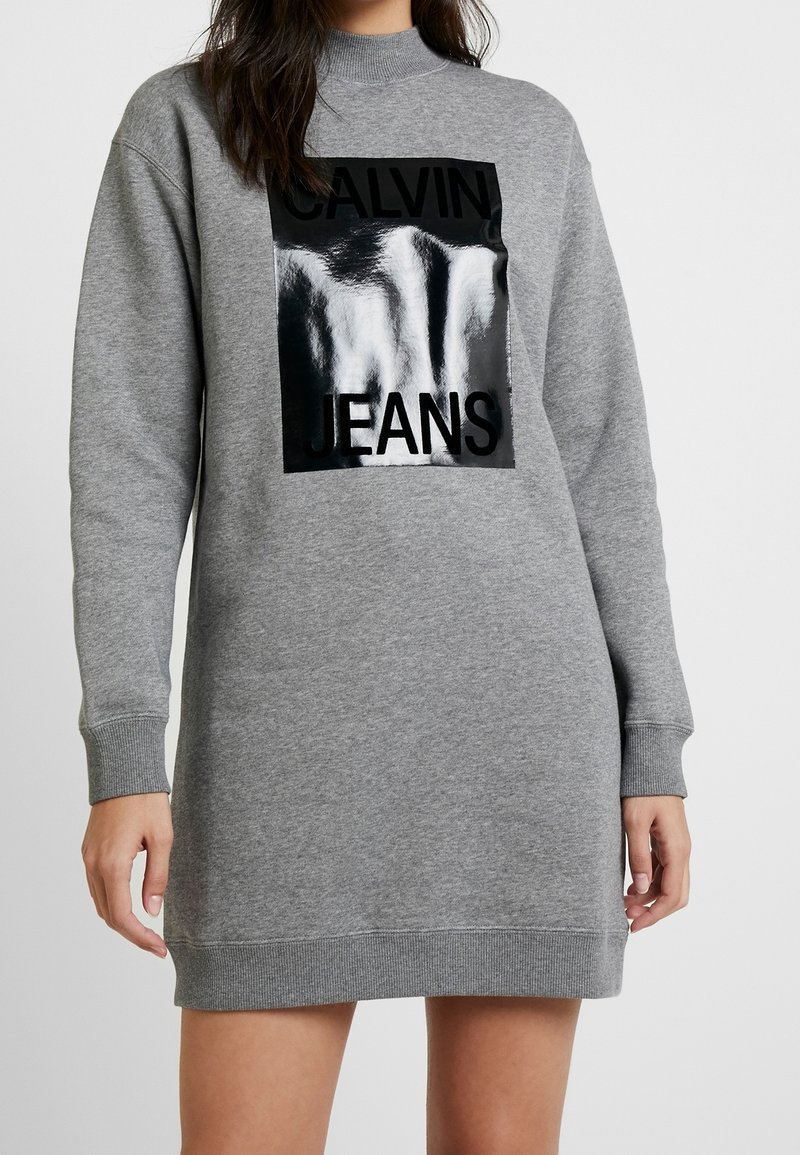 Vestido sudadera gris con mangas largas, puños y dobladillo acanalados. Presenta un gráfico rectangular negro brillante con el texto "CALVIN JEANS".