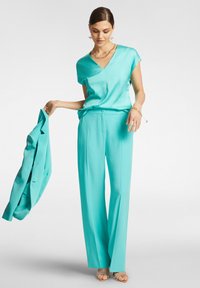 Top in satin turchese con scollatura a V e maniche corte, abbinato a pantaloni larghi coordinati. Il modello tiene in mano un blazer abbinato.