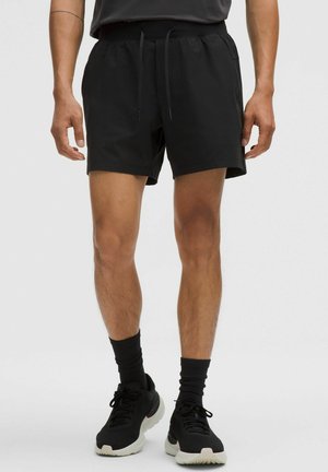 Man die zwarte sportkorte broek, zwarte sokken en zwarte sneakers draagt, staand tegen een effen lichte achtergrond.