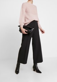 Kobieta w różowym swetrze z dzianiny, czarnych szerokich spodniach, czarnych botkach na obcasie, trzymająca czarną skórzaną torebkę kopertową.