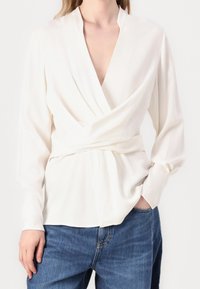 Biała bluzka z długimi rękawami, głębokim dekoltem w serek i drapowanym przodem. Gładki materiał i detal w talii. Noszona z niebieskimi dżinsami.