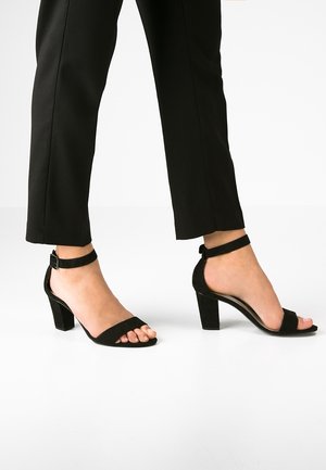 Pieds portant des sandales noires à talon bloc et bride cheville, à bout ouvert, associées à un pantalon noir court sur fond blanc.