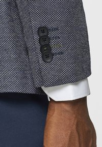 Blazer a maniche con motivo blu navy e bianco, dotato di quattro bottoni neri e polsino bianco a contrasto. Tessuto testurizzato con una vestibilità sartoriale.